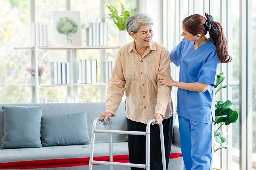Asian young professional successful friendly female nurse in blue hospital uniform helping supporting physical therapy senior old pensioner unhealthy injury woman patient walking via four legs walker.